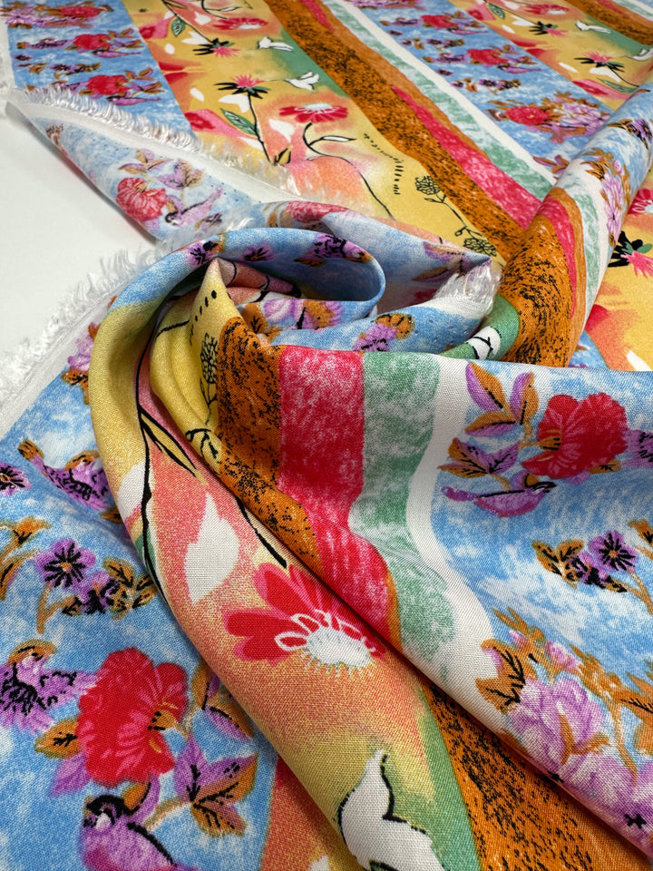 Designer Rayon - Wildframe by Super Cheap Fabrics is a vibrant patterned fabric (145cm wide) with bold stripes, flowers, and abstract shapes in red, yellow, blue, pink, and green tones. Shown draped and partially rolled on a white surface.