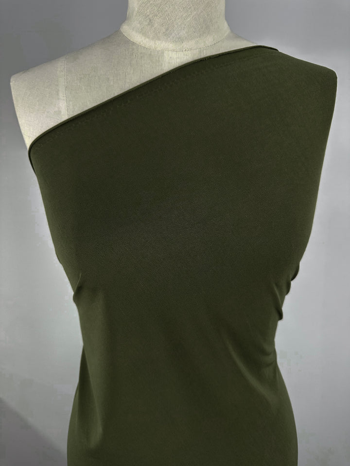 A mannequin showcases an olive green, one-shoulder top made from Super Cheap Fabrics' ITY Knit (150cm), featuring smooth, fitted, medium-weight material that stretches across the torso while exposing one shoulder and part of the neckline.