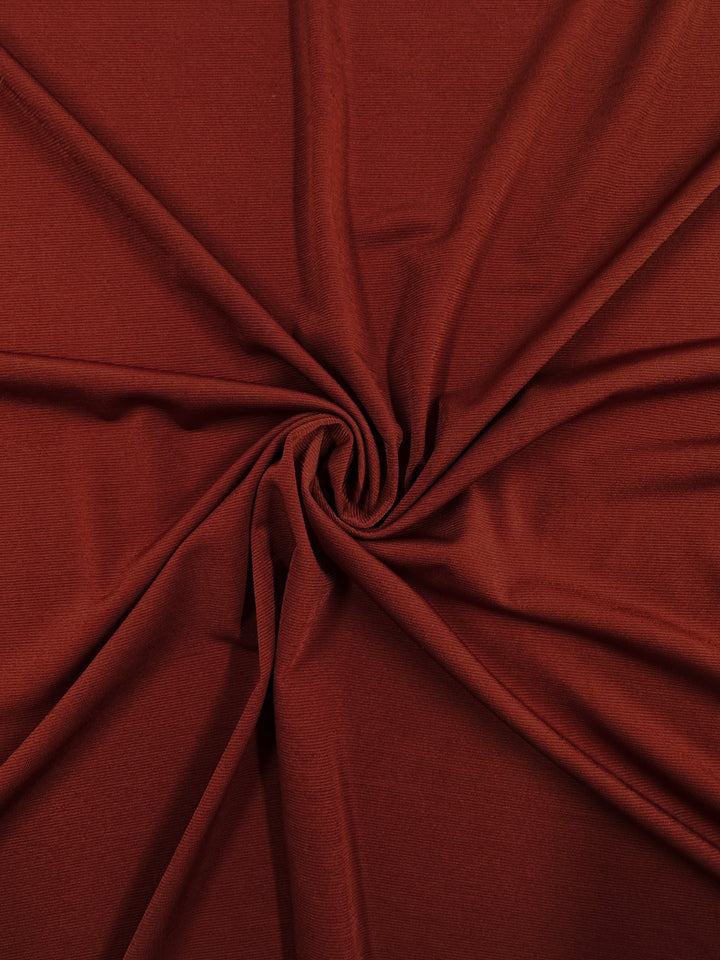 Close-up of Super Cheap Fabrics’ ITY Knit - Ketchup (150cm), a rust-colored polyester spandex with a soft texture, shown gathered and twisted at the center to create gentle folds and a circular pattern.