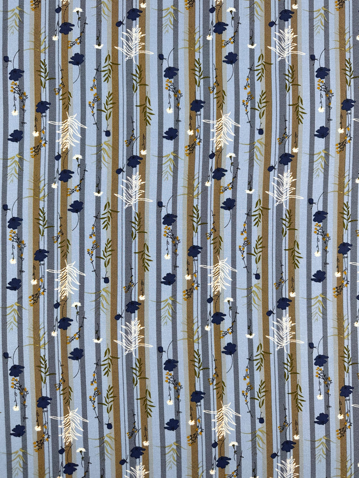 Super Cheap Fabrics’ Designer Rayon - Fieldstripe Bloom (145cm) features blue rayon with vertical brown and light blue stripes, accented by scattered navy and white floral and branch motifs for a woodland-inspired look.