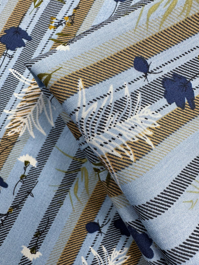 Designer Rayon - Fieldstripe Bloom by Super Cheap Fabrics features blue rayon with diagonal brown, white, and black stripes, plus a white and navy flower and leaf pattern. Slightly folded to show texture. Width: 145cm.
