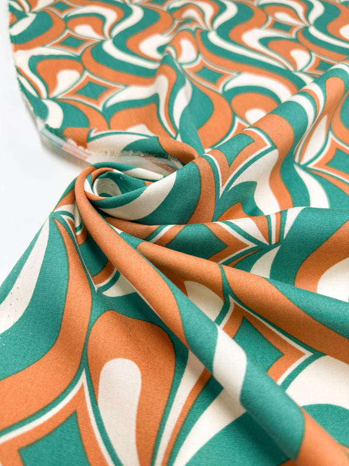 A close-up of Super Cheap Fabrics’ Designer Rayon - Retro Pulse (145cm) shows bold orange, green, and white geometric wavy and circular patterns on soft fabric, slightly gathered to highlight its smooth texture and vibrant look.