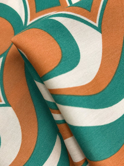 A close-up of Super Cheap Fabrics' Designer Rayon - Retro Pulse - 145cm reveals its bold teal, orange, and white retro pattern, with vibrant curved shapes and lines highlighted on the softly folded material.