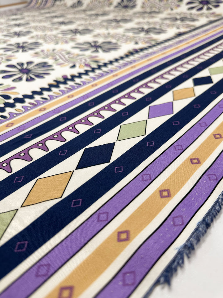A close-up of Super Cheap Fabrics' Designer Rayon - Lilac Tempo (145cm) shows geometric and floral patterns in navy, purple, gold, cream, and green with rows of diamonds and ornate edging.