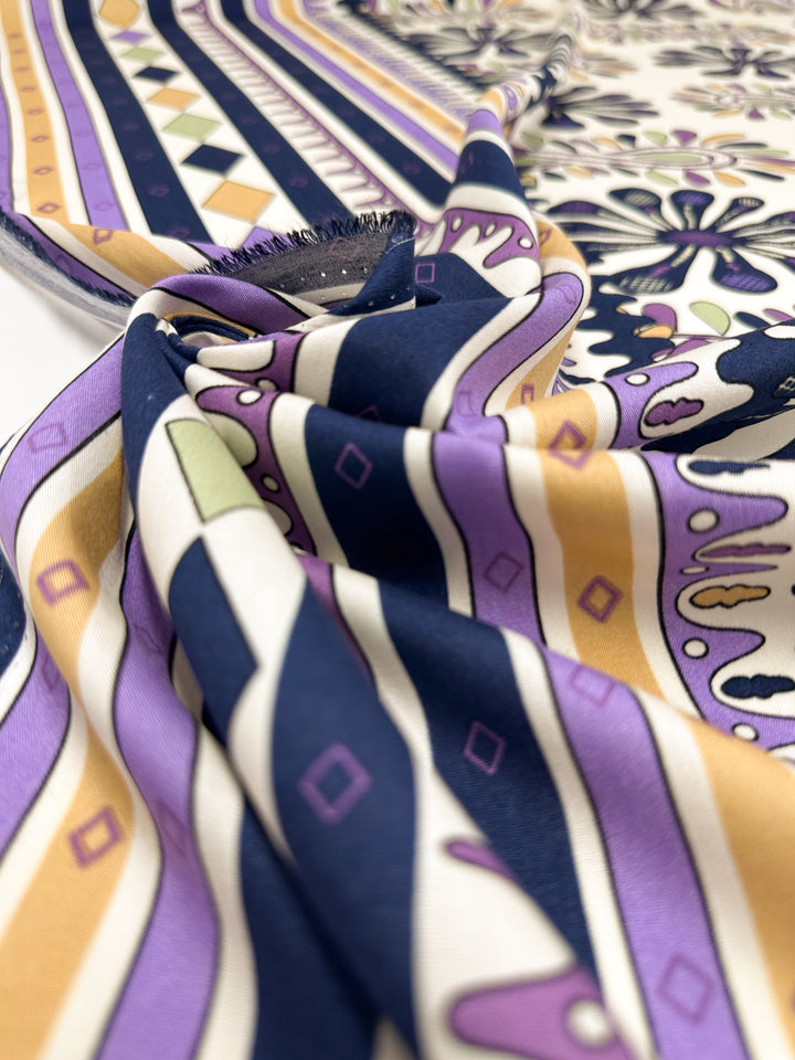 A close-up of Super Cheap Fabrics' Designer Rayon - Lilac Tempo (145cm), showing its smooth, silky texture and colorful pattern of stripes and geometric shapes in purple, navy, yellow, cream, and pale green, loosely folded and draped.