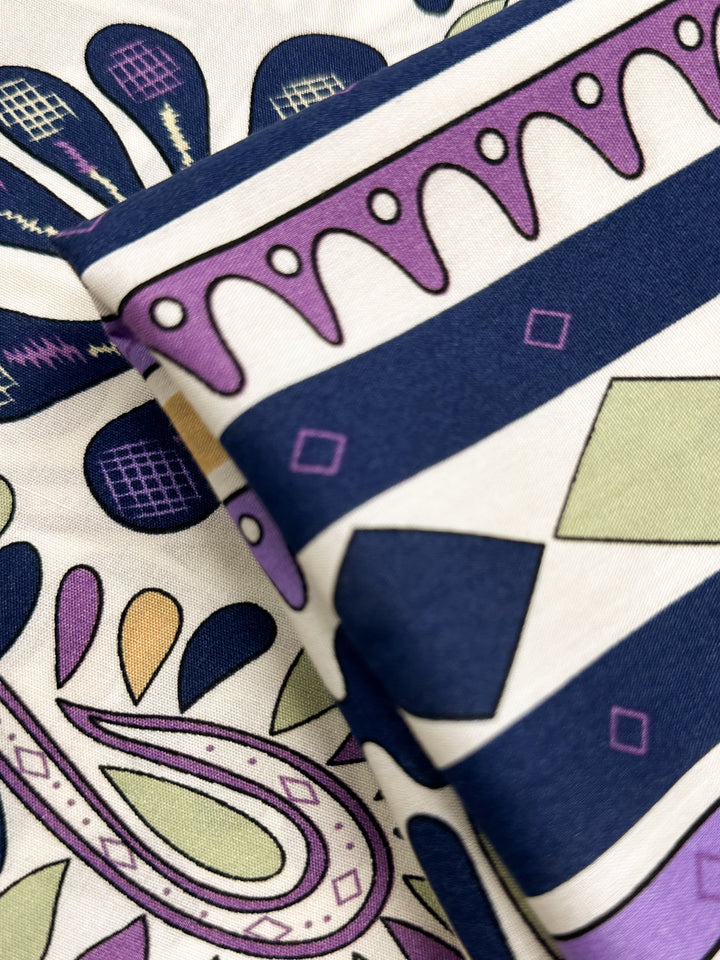 A close-up of Super Cheap Fabrics’ Designer Rayon - Lilac Tempo (145cm), slightly folded, showing vibrant navy blue, purple, green, and yellow geometric and paisley patterns with stripes, diamonds, teardrops, and curves.