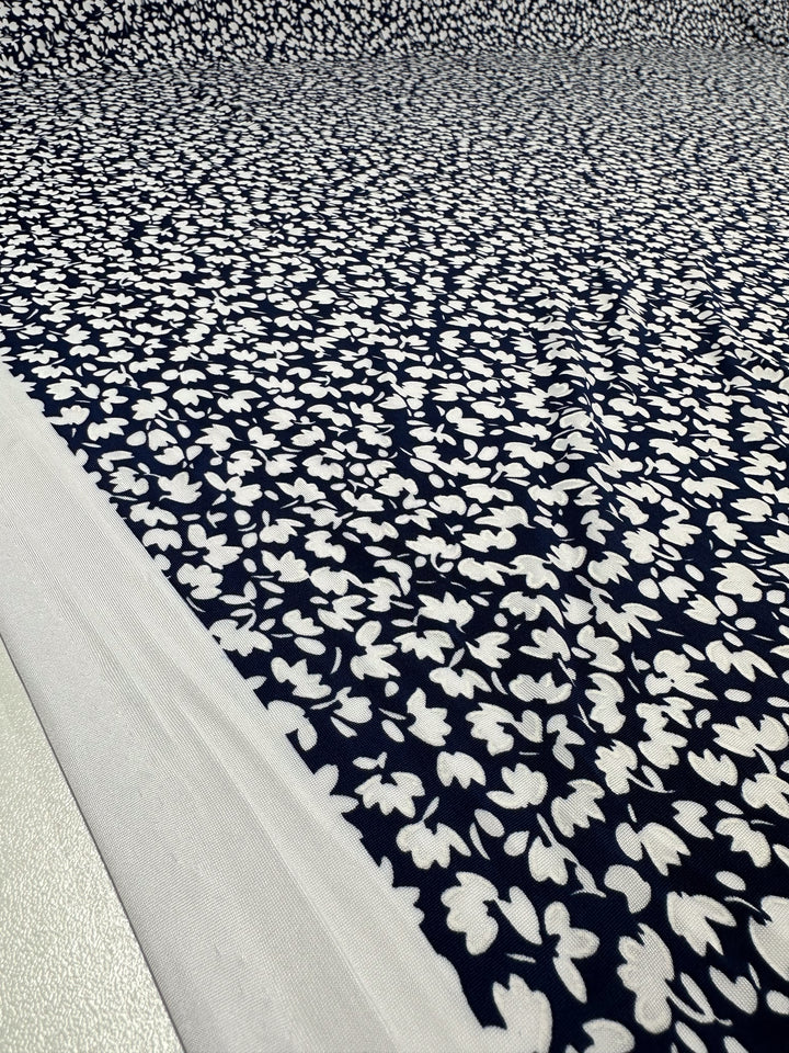 A close-up of Super Cheap Fabrics' Textured Lycra - Night Bloom Micro (150cm), featuring a navy blue base with dense small white floral prints, laid flat with a plain white border along the edge.