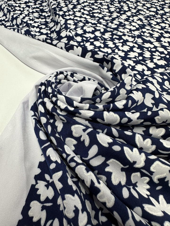 A close-up of Super Cheap Fabrics' Textured Lycra - Night Bloom Micro (150cm), a navy blue medium weight lycra with a white floral pattern, partially rolled and displayed on a flat surface with white background edges.