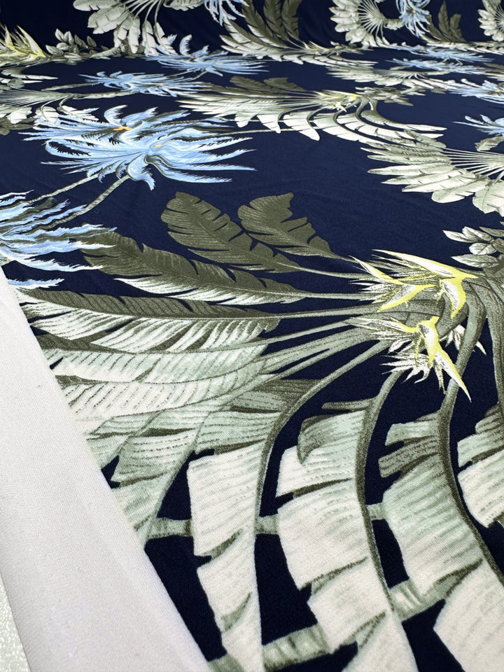 Close-up of Super Cheap Fabrics’ Printed Lycra - Islander Drift (150cm), showing stretch polyester spandex with a tropical leaf and flower pattern in green, blue, and white on dark blue. Fabric edge appears in the lower left corner.