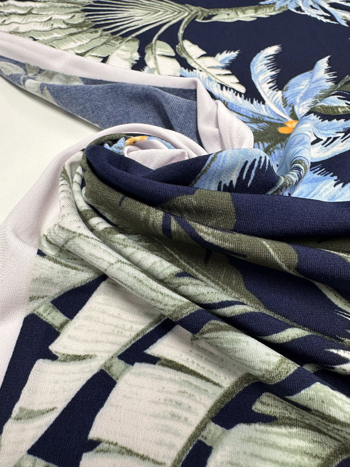 Close-up of Super Cheap Fabrics' Printed Lycra - Islander Drift (150cm), a soft stretch fabric in blue, green, white, and yellow tropical leaf print, gathered into a swirl on a flat surface.