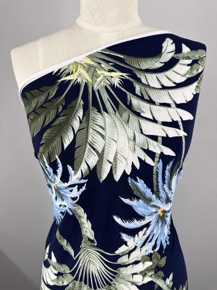 A dress form is draped in Super Cheap Fabrics' Printed Lycra - Islander Drift (150cm), featuring navy blue stretch fabric with a bold tropical leaf and flower pattern in green, white, and pale blue, wrapped asymmetrically over one shoulder.