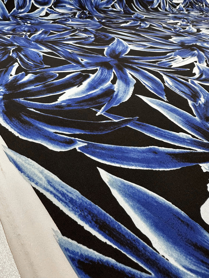 Close-up of Super Cheap Fabrics' Printed Lycra - Cobalt Charge (150cm): a medium weight lycra with a bold, abstract blue and white floral pattern on a black base, featuring dynamic overlapping leaves and detailed brush-like strokes.