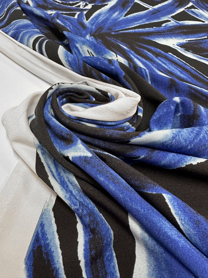 A close-up of Super Cheap Fabrics’ Printed Lycra - Cobalt Charge (150cm), featuring a blue, black, and white abstract leaf pattern. The fabric’s medium weight texture and spiral folds are clearly visible on a flat surface.