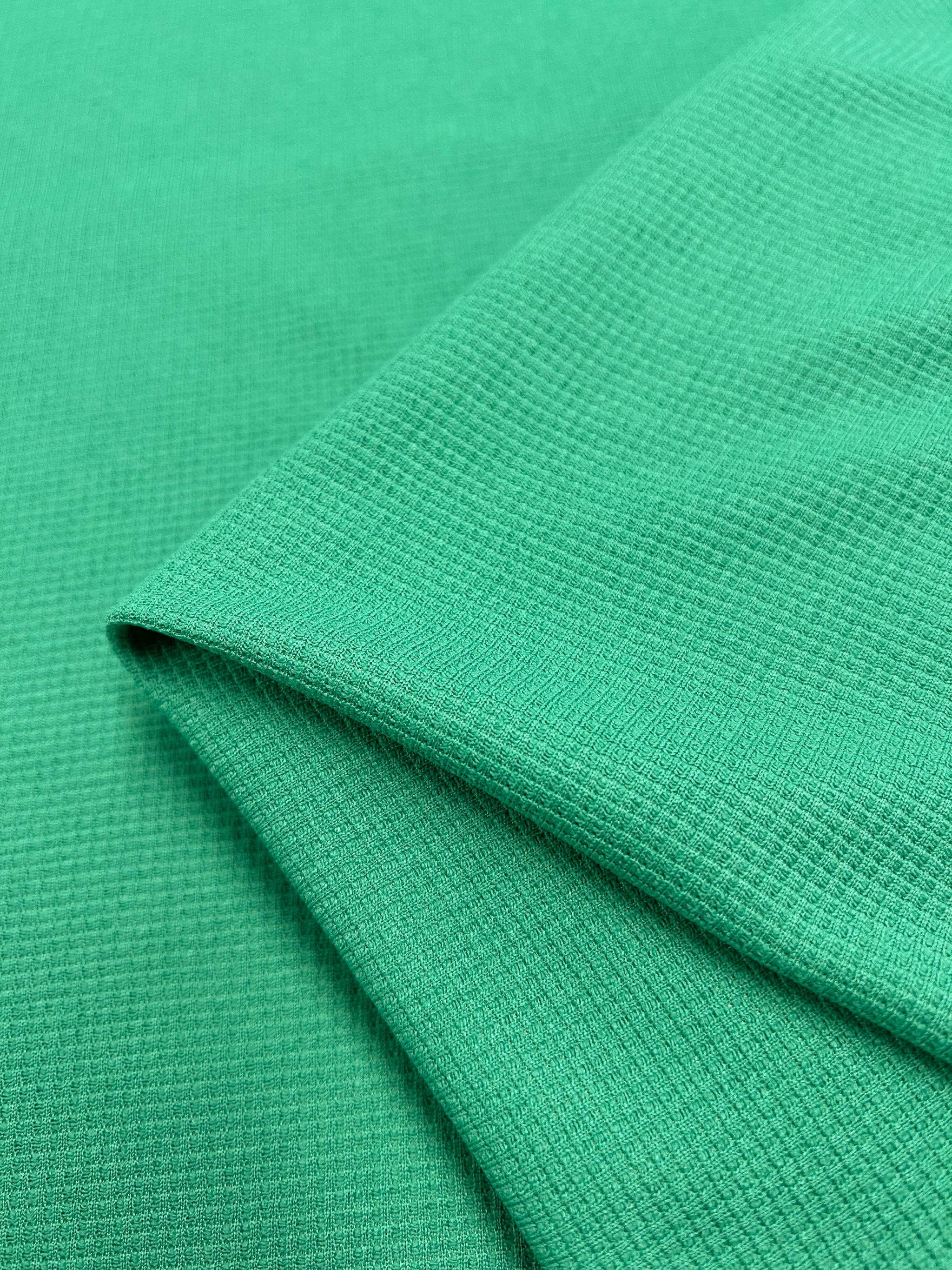 A close-up of an emerald green, honeycomb fabric with a folded corner, revealing its fine grid-like pattern and smooth surface. The fabric appears lightweight and soft, suitable for clothing or other textile applications. This is the Waffle Knit - Jade Cream - 170cm from Super Cheap Fabrics.
