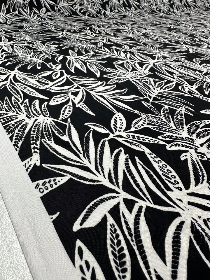 Close-up of Super Cheap Fabrics’ Textured Lycra - Shadow Fern (150cm) in black, featuring a detailed white fern and foliage print with dense overlap and a bold white border along the left edge.
