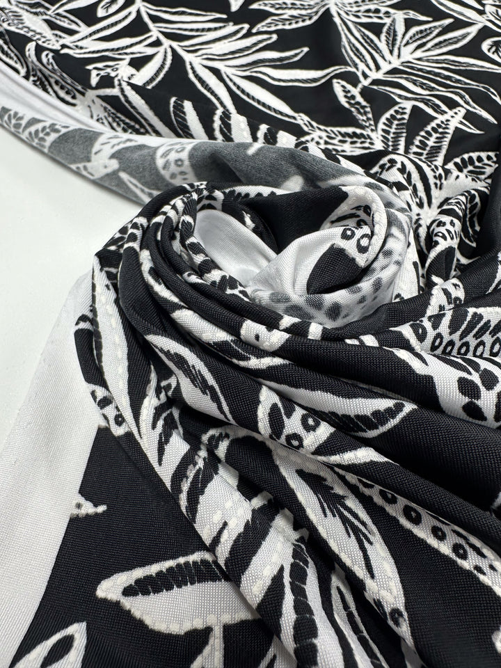 Textured Lycra - Shadow Fern by Super Cheap Fabrics features a bold black and white botanical leaf pattern with intricate details, shown partially rolled and folded on a flat surface. Width: 150cm.