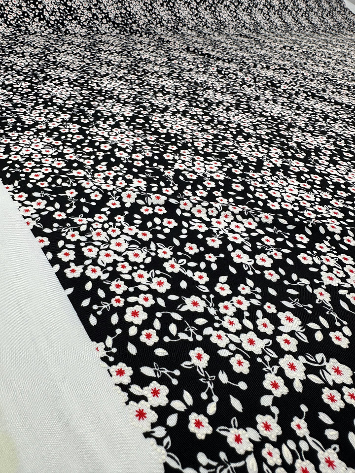 A close-up of Super Cheap Fabrics' Textured Lycra - Sugar Pollen (150cm), showing medium-weight lycra with a black base and a repeating pattern of small white flowers with red centers, laid flat with a white border on the edge.