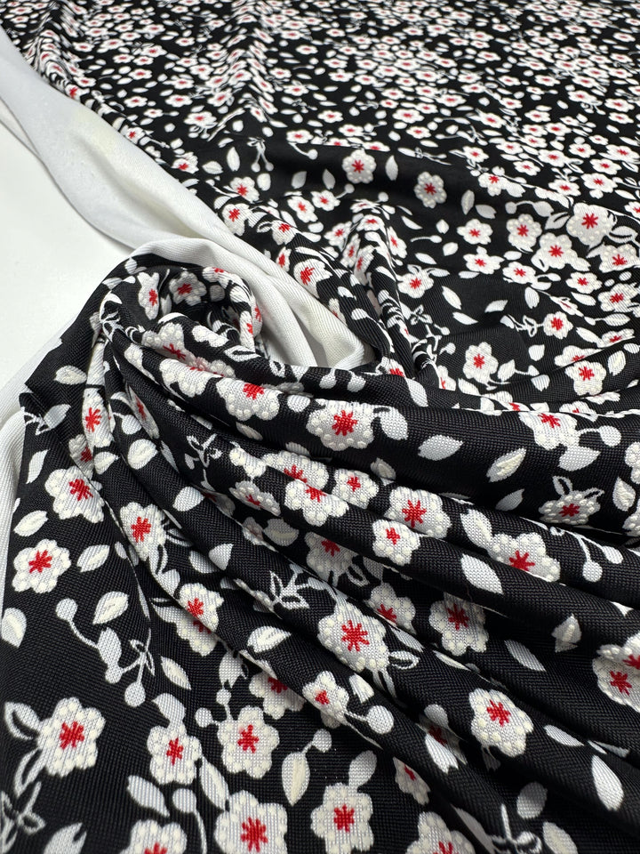 A close-up of Super Cheap Fabrics’ Textured Lycra - Sugar Pollen (150cm) shows its white and red floral pattern on black, with folds revealing the medium-weight texture and the print’s detail on both front and reverse sides.