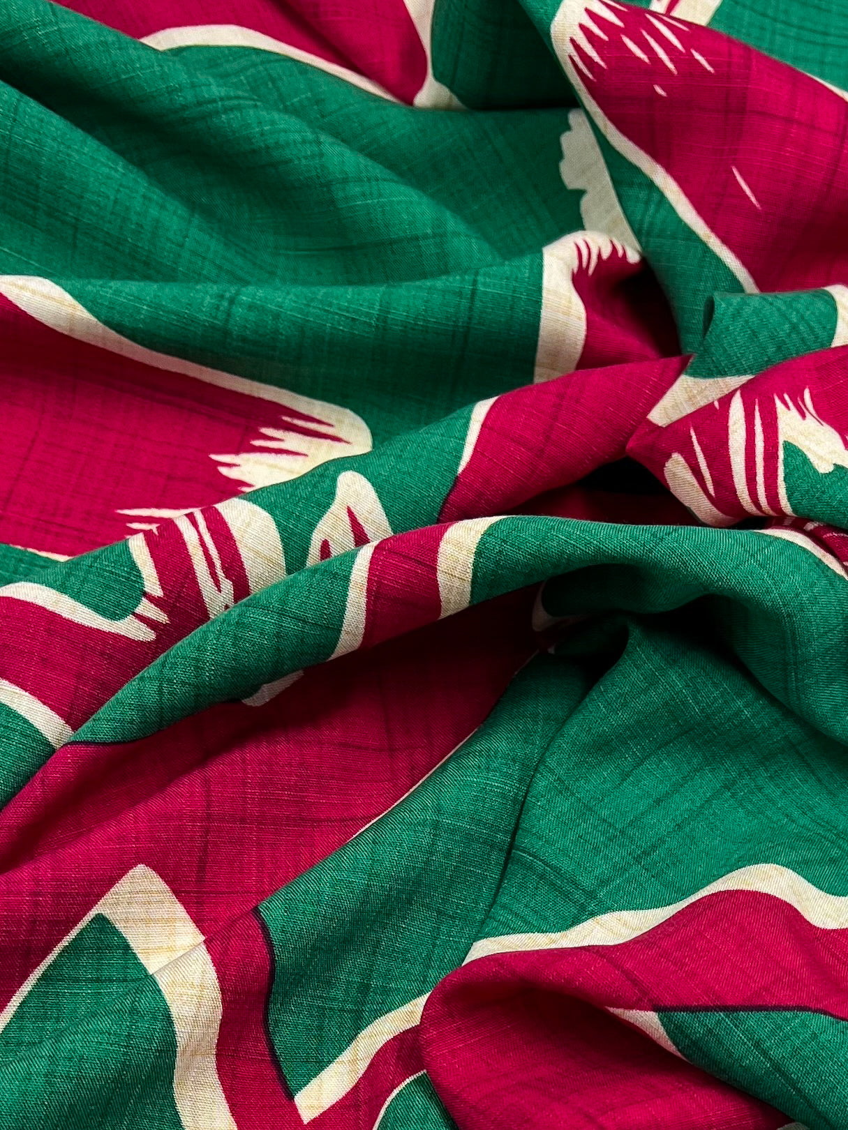 A close-up of a folded lightweight fabric featuring a vibrant pattern. The Bamboo Rayon - Dance Pepper Green - 150cm by Super Cheap Fabrics has a green base with red and white abstract shapes and lines. The texture is slightly crinkled, and the weave of this beginner-friendly sewing material is visible.
