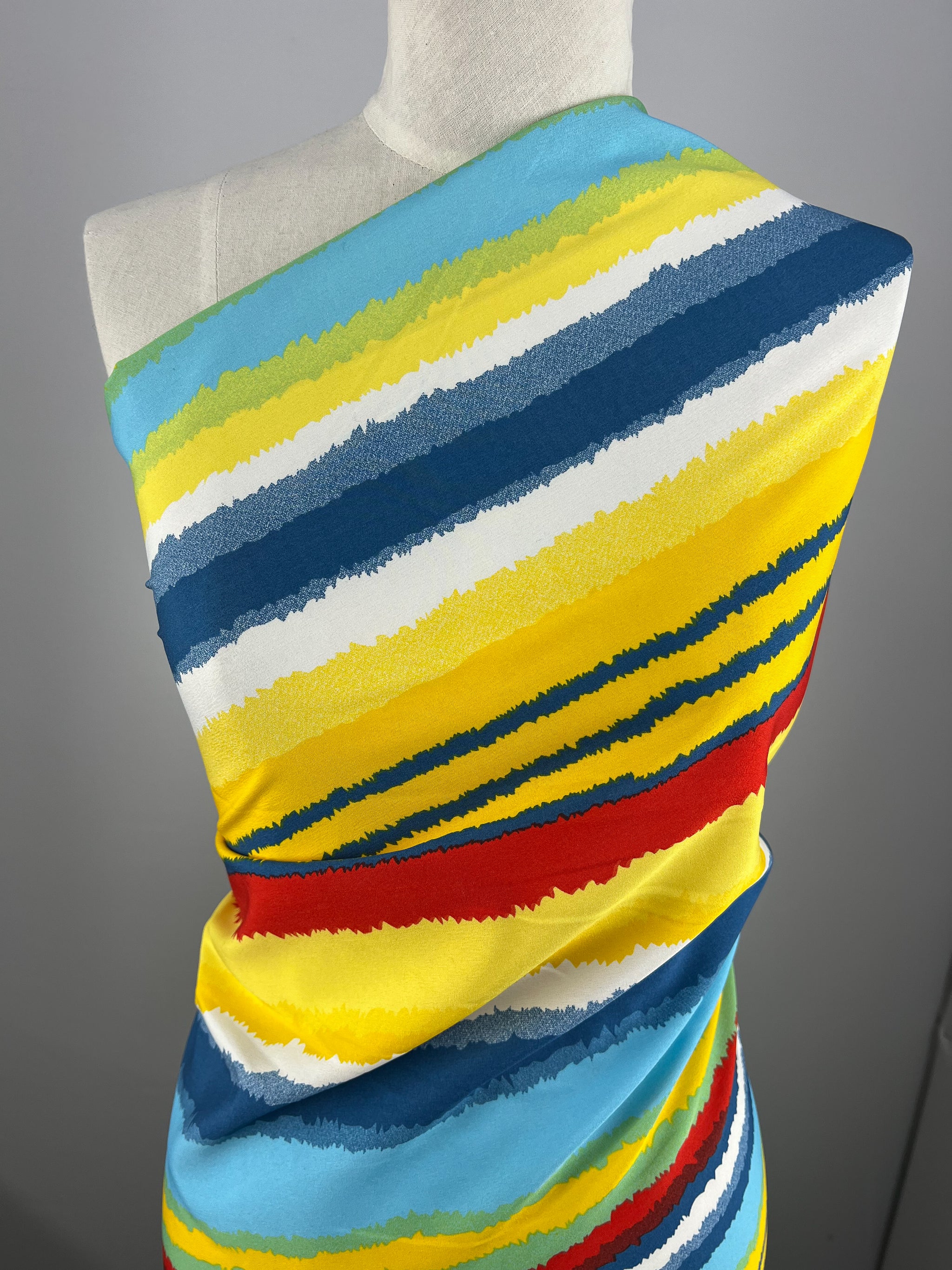 A white mannequin is draped with a colorful, versatile polyester fabric featuring bold horizontal stripes in blue, yellow, red, white, and light blue. The Deluxe Print - Beachside - 155cm by Super Cheap Fabrics wraps around the torso, showcasing the vibrant design. The background is plain and gray.