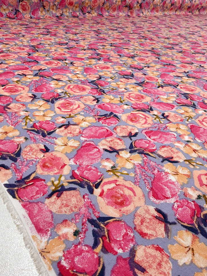 A close-up of Designer Rayon - Botanic Crush (142cm) by Super Cheap Fabrics displays a dense, colorful floral pattern with large pink and peach roses and green leaves on a light purple background, highlighting its vibrant, versatile design.