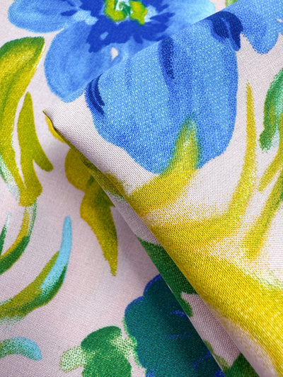 A close-up of Super Cheap Fabrics' Designer Rayon - Bluebell Blush (145cm), showcasing its vibrant blue and green floral print with yellow accents on a light background, and subtle folds that highlight the fabric’s soft texture.