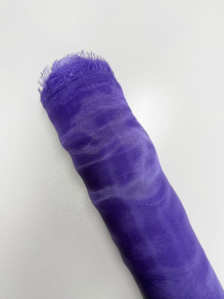 A rolled-up piece of Super Cheap Fabrics’ Organza - Sand Verbena (150cm), sheer purple with frayed edges, lies on a smooth white surface.