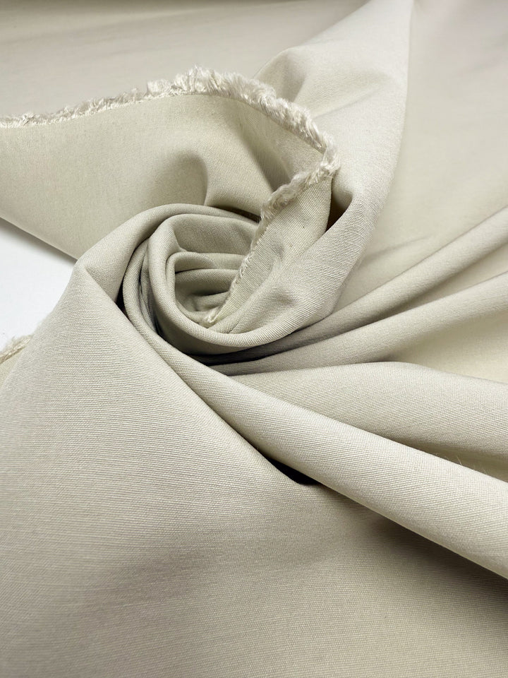 Super Cheap Fabrics' Cotton Bengaline in Bone White (150cm) is displayed with a slightly rough, unfinished edge and artfully twisted into a spiral, highlighting its smooth texture and soft look.