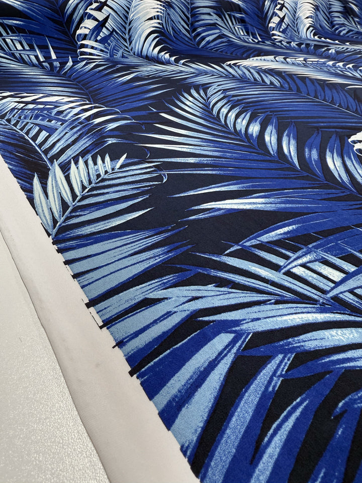 A close-up of Super Cheap Fabrics' Printed Lycra - Sapphire Jungle (150cm), showcasing a blue and white palm leaf pattern on a dark background, with the fabric edge visible against a light surface.