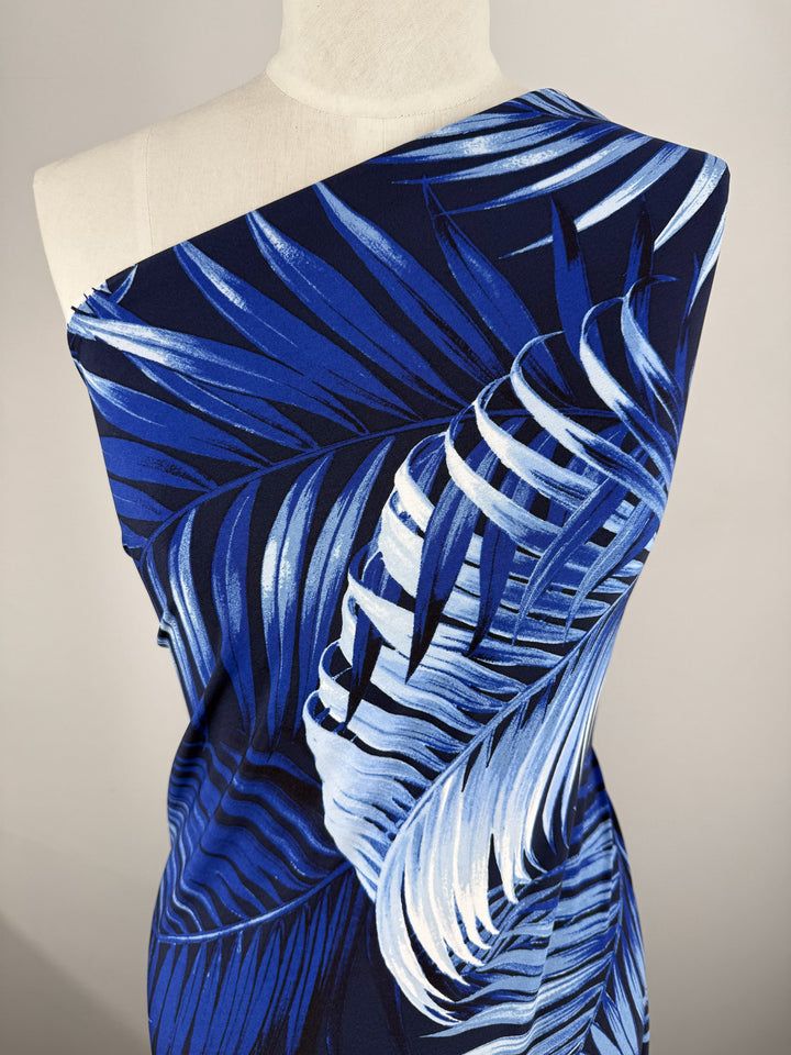 A dress form displays Super Cheap Fabrics' Printed Lycra - Sapphire Jungle (150cm), a medium-weight fabric with a bold blue and white tropical palm leaf print, draped off one shoulder against a neutral background.