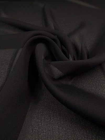 Close-up of Super Cheap Fabrics' Plain Georgette - Black (114cm), showcasing its sheer black polyester with a soft, lightly textured surface. The fabric is gently twisted, forming delicate folds and a swirling pattern.