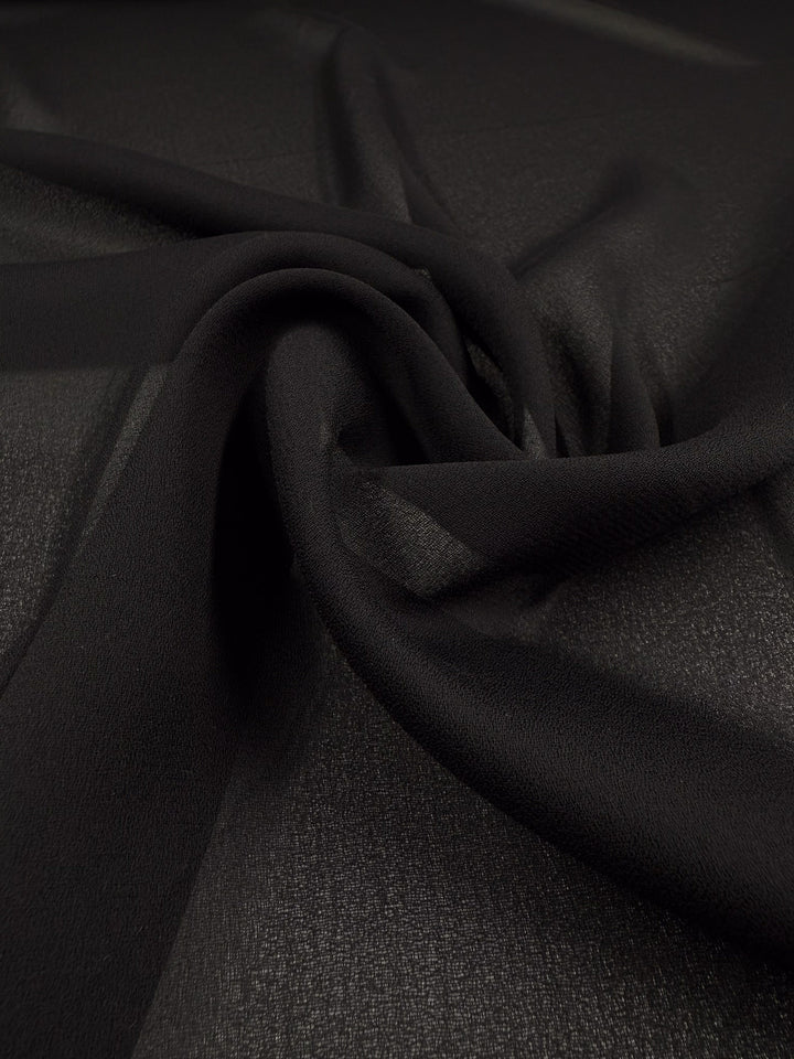 Close-up of Super Cheap Fabrics' Plain Georgette - Black (114cm), showcasing its sheer black polyester with a soft, lightly textured surface. The fabric is gently twisted, forming delicate folds and a swirling pattern.