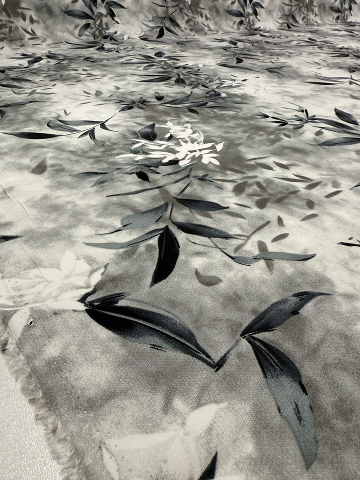 A close-up of Super Cheap Fabrics' Designer Rayon - Misted Olive (147cm), featuring a soft, lightweight fabric in gray and white with subtle floral and leaf motifs in black, dark gray, and white, finished with a watercolor-like effect.