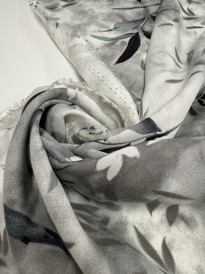 A close-up of Super Cheap Fabrics' Designer Rayon - Misted Olive (147cm), a soft and lightweight rayon fabric with a subtle floral pattern in gray, white, and black, gently folded on a white surface.