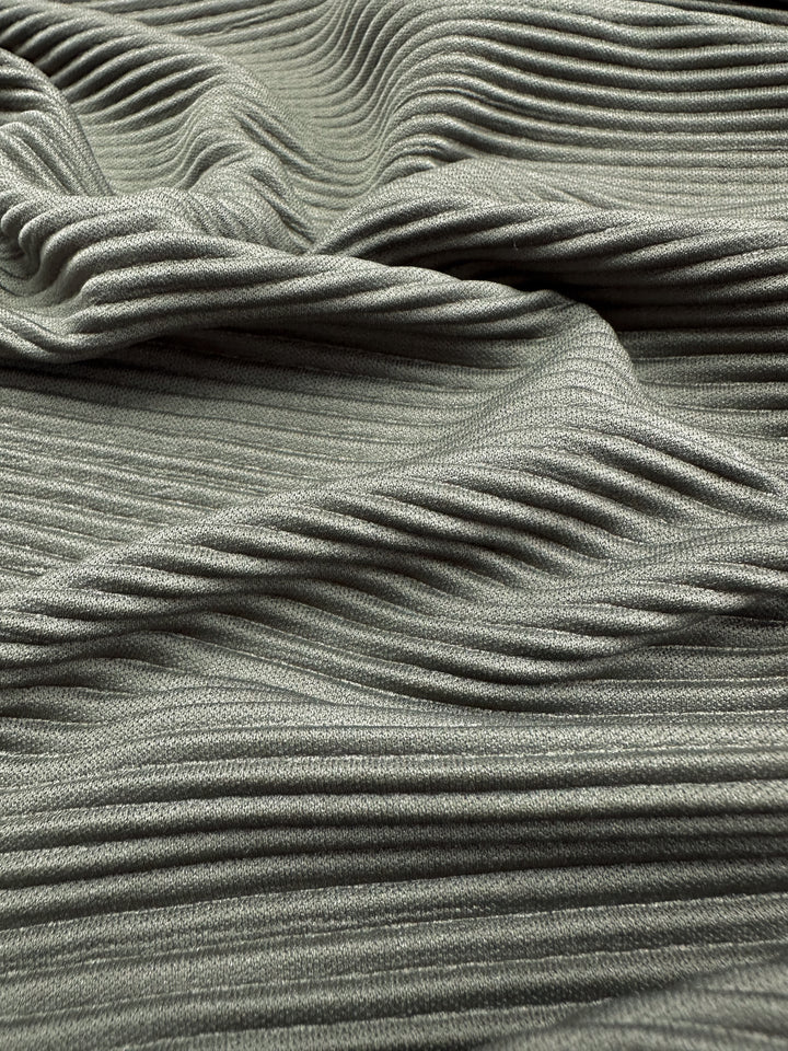 A close-up of Super Cheap Fabrics' Pleat - Pewter - 140cm showcases a lightweight, sage green polyester with fine horizontal ribbing. Draped in gentle folds, the fabric creates soft shadows and conveys a sense of movement.