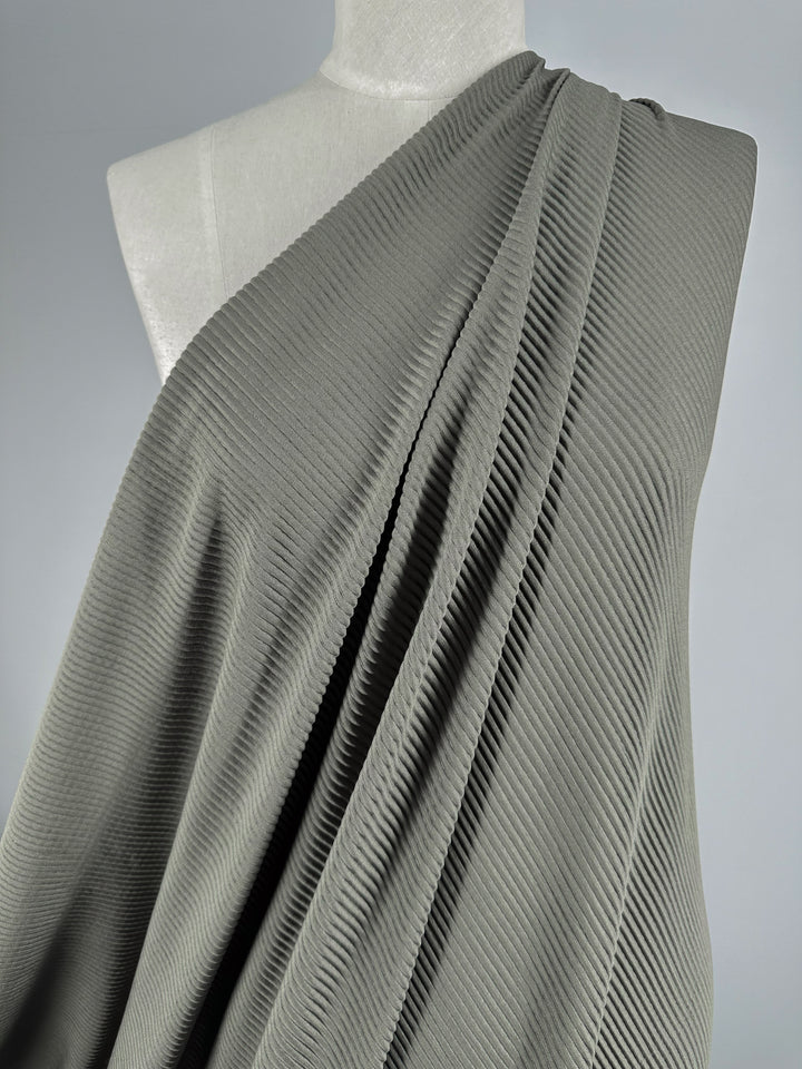 A close-up showcases the Pleat - Pewter - 140cm fabric by Super Cheap Fabrics, featuring lightweight polyester with a ribbed texture. Draped over a mannequin's shoulder against a plain background, it forms gentle folds that highlight its vertical texture.
