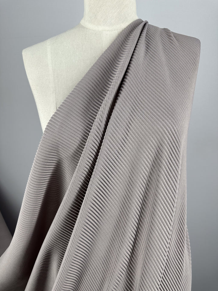 The Super Cheap Fabrics' Pleat in Violet Ice, a light gray textured polyester with a pleated pattern, is elegantly draped over a mannequin against a neutral gray background.