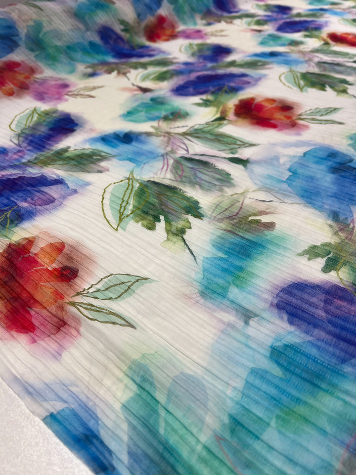 The Textured Tencel - Dream Flor by Super Cheap Fabrics features a watercolor-style floral pattern with blue, red, and purple flowers on textured fabric. Green leaves are interspersed in the design, offering an artistic touch that combines vivid charm with sustainability.