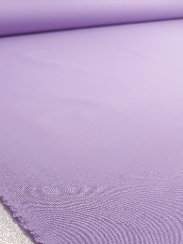 A close-up of Super Cheap Fabrics' Milano Ponte - Viola (160cm) shows the smooth, light purple stretch fabric laid flat with a slightly frayed hem at the lower left and a roll of heavy weight ponte in the background.