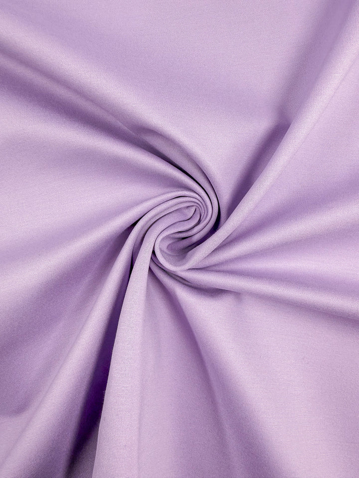 A close-up of Milano Ponte - Viola by Super Cheap Fabrics shows lavender heavyweight stretch fabric, 160cm wide, with a smooth texture gathered and twisted at the center to create soft spiral folds.