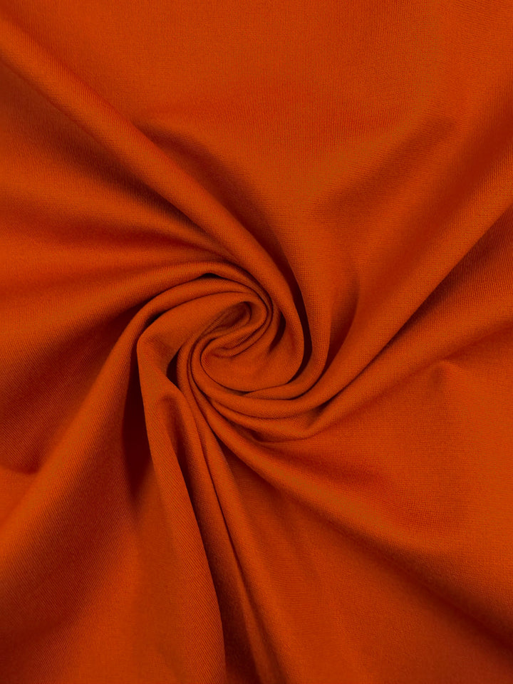 Close-up of "Milano Ponte - Chili - 160cm" by Super Cheap Fabrics, featuring soft orange ponte fabric arranged in a swirl, with folds and shadows adding depth and movement.