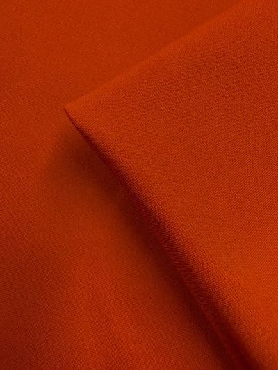 A close-up of Super Cheap Fabrics' Milano Ponte - Chili (160cm) shows the orange heavyweight fabric, with one piece folded diagonally over another to highlight its texture and weave.