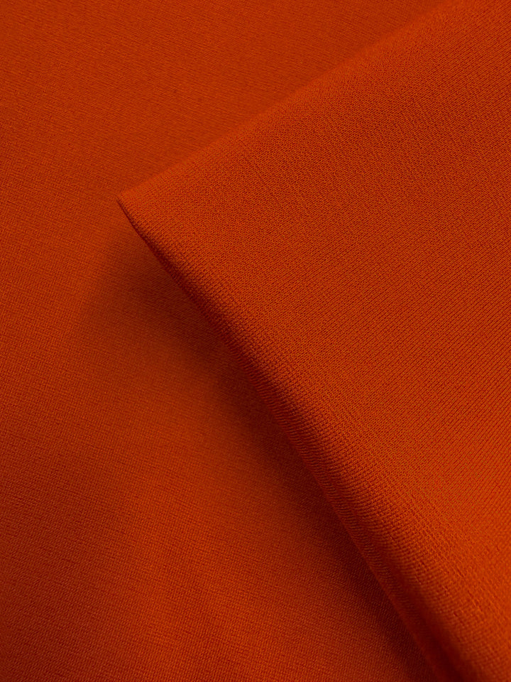 A close-up of Super Cheap Fabrics' Milano Ponte - Chili (160cm) shows the orange heavyweight fabric, with one piece folded diagonally over another to highlight its texture and weave.