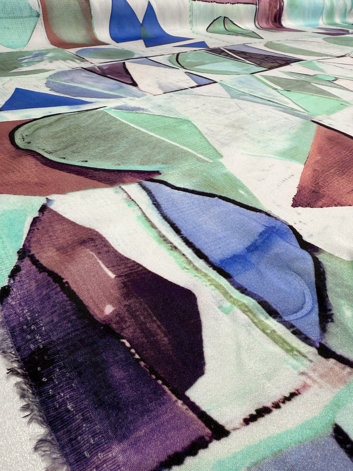 A close-up of Super Cheap Fabrics’ Mercerised Rayon - Mid Mod Mirage (150cm), featuring breathable, lightweight rayon with abstract geometric shapes in teal, blue, purple, and burgundy on a soft textured surface.