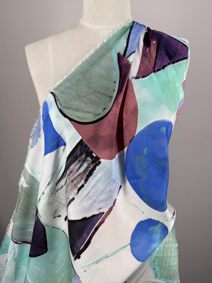 A mannequin displays "Mercerised Rayon - Mid Mod Mirage - 150cm" by Super Cheap Fabrics, a lightweight, breathable fabric with a soft texture and abstract blue, maroon, mint green, and black patterns on white. Edges are unfinished.
