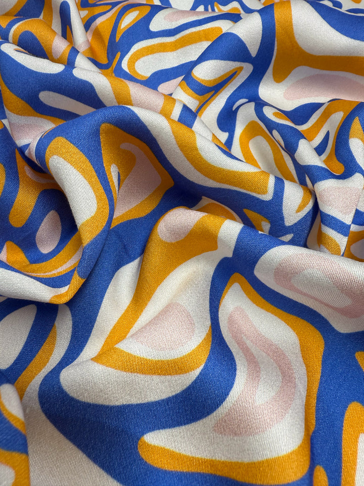 A close-up of Super Cheap Fabrics' Mercerised Rayon - Electric Ember (145cm), showing its lightweight, breathable drape and bold abstract pattern in blue, yellow, white, and pale pink with fluid, wavy shapes.