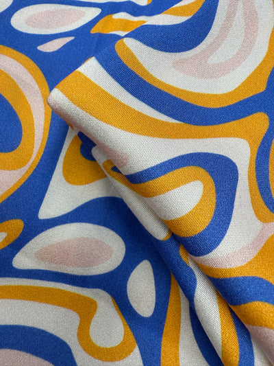 A close-up of Super Cheap Fabrics' Mercerised Rayon - Electric Ember (145cm), showcasing lightweight, breathable rayon with a bold abstract pattern in blue, yellow, pink, and white plus a subtle sheen and fluid drape.