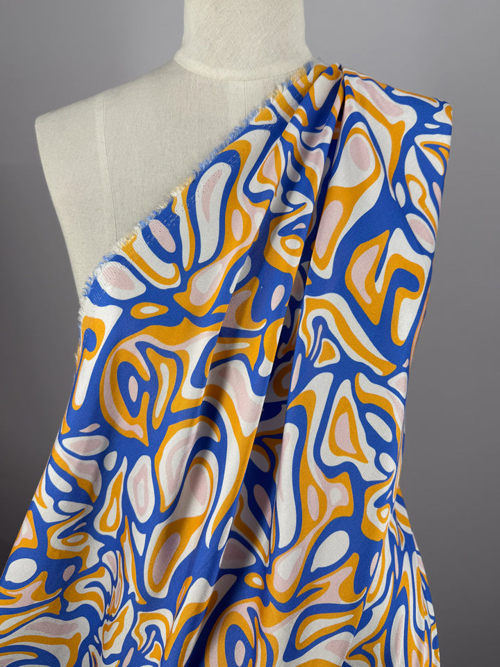 A mannequin displays Super Cheap Fabrics' Mercerised Rayon - Electric Ember (145cm), featuring a lightweight, boldly patterned fabric in blue, yellow, white, and pink with swirling shapes, draped over one shoulder against a plain gray background.
