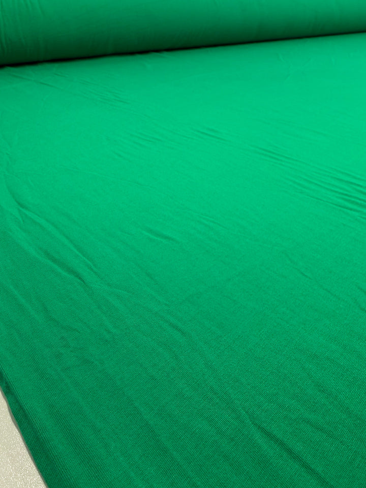 A close-up of Super Cheap Fabrics' Cotton Jersey - Parakeet - 165cm, a bright green 100% cotton fabric roll, laid flat to show its subtle creases and smooth texture.