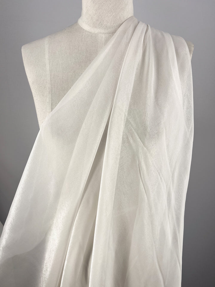 Super Cheap Fabrics’ Silk Mousseline - Alabaster - 135cm, a white, extra lightweight sheer silk, is draped over a dress form to create soft flowing folds that reveal its texture and subtle translucence against a plain gray backdrop.